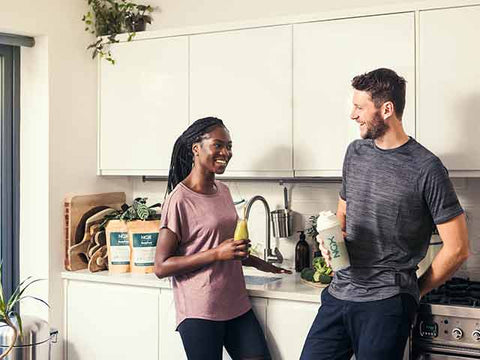 Woman in pink top and man in grey sports top standing next to one another. The women is smiling holding a smoothie, and the man is holding a shaker.